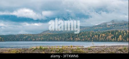 Nordeuropäische Herbstlandschaft mit einem ruhigen See, der die bewölkte Himmel reflektiert, mit Hügeln, die mit Kiefern und Laubbäumen bedeckt sind, die ein lebhaftes Herbstlaub zeigen Stockfoto