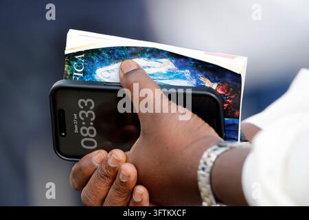 Eine Nahaufnahme von den Händen einer Person, die ein Smartphone mit der Uhrzeit und ein illustriertes religiöses Heft während der Messe des Papstes hält. Stockfoto