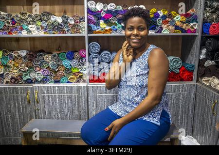 Ein lächelnder ruandischer Ladenbesitzer sitzt vor Regalen, die mit Rollen farbenfroher Stoffe gefüllt sind, in Ruanda. Stockfoto