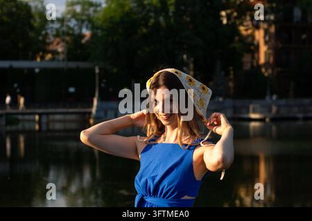 Porträt einer Frau bei Sonnenuntergang in einem grünen Park, die ein tiefblaues Kleid und einen gestrickten kleinen Schal auf dem Kopf trägt, der die warme Sommeratmosphäre und Freiheit illustriert Stockfoto