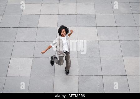 Lächelnder junger schwarzer Mann, der auf einem betonierten urbanen Platz mit Fliesen springt und tanzt Stockfoto