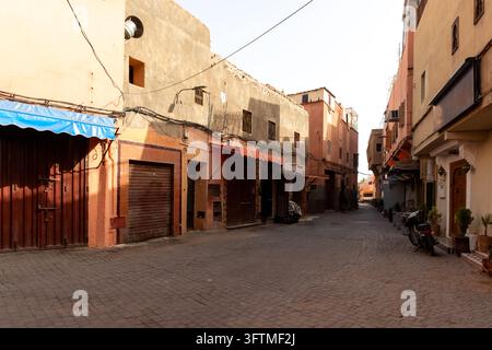 Ruhige Kopfsteinpflasterstraße in der Medina von Marrakesch, gesäumt von verwitterten Terrakotta-Gebäuden, geschlossenen Ladenfronten, bunten Markisen, Überkopfkabeln und warmer Stockfoto