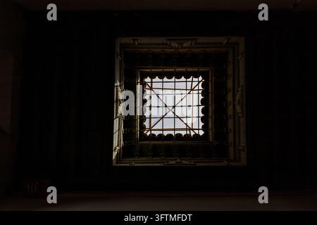 Eine symmetrische Aufnahme, die sich auf ein einzelnes gittermusteriges Fenster mit eisernen Sicherheitsgittern konzentriert, das in eine dunkle Decke eingelassen ist und Tageslicht in Marokko hereinlässt.... Stockfoto