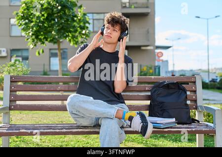 Entspannter junger Mann, Student, der auf einer Bank im Stadtpark sitzt, mit kabellosen Over-Ear-Kopfhörern berührt und Musik oder Podcast hört. Freizeit-Audiobo Stockfoto
