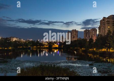 Lichter hoher Wohnhäuser spiegeln sich in einem See mit Seerosen und Schilf bei Nacht im Bezirk Poznyaky von Kiew Stockfoto