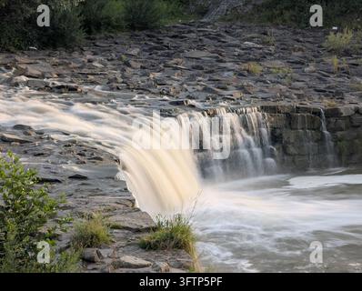 Ein Wasserfall stürzt über felsiges Gelände und erzeugt einen sanften Fluss, während er in ein Becken darunter stürzt. Um den Wasserfall herum befinden sich Flecken von üppigem grünen V Stockfoto