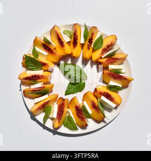 Pfirsichsalat mit Mozzarellakäse, angeordnet in einem kreisförmigen Design auf einem weißen Teller mit frischen Basilikumblättern, isoliert auf weißem Hintergrund Stockfoto