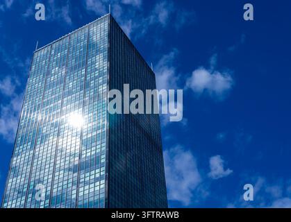 Niedrigwinkelaufnahme des Treptowers-Büroturms in Berlin Alt-Treptow, wobei das helle Sonnenlicht auf seiner Glasfassade reflektiert wird vor einem blauen, bewölkten Himmel. Stockfoto