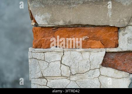 Nahaufnahme-Makrofotografie einer verwitterten Architekturwand mit altem, rissigem grauem Zementputz, der rohe rote Ziegelsteine darunter freilegt Stockfoto