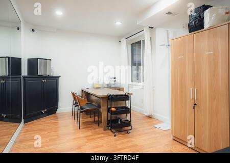 Kleines Büro oder Arbeitszimmer mit Holzschreibtisch und zwei Mid-Century-Modern-Stühlen neben dem Fenster mit weißen Vorhängen, schwarzer dreistufiger Rollwagen im Vordergrund Stockfoto