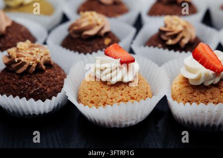 Nahaufnahme von verschiedenen Mini-Cupcakes in weißen Papierförmchen mit gepipster Sahneglasur und frischen Erdbeerscheiben auf krümeligen goldenen und Schokoladenböden arr Stockfoto