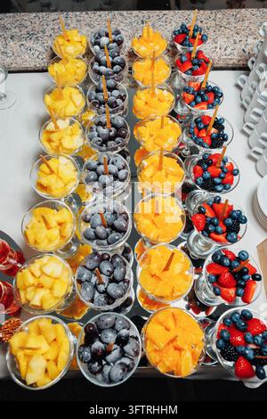 Reihen von Dessertgläsern gefüllt mit Ananas, Trauben, Orangen und gemischten Beeren auf spiegelnder Buffetpräsentation mit Holzspießen zum Servieren am Veranstaltungstisch Stockfoto