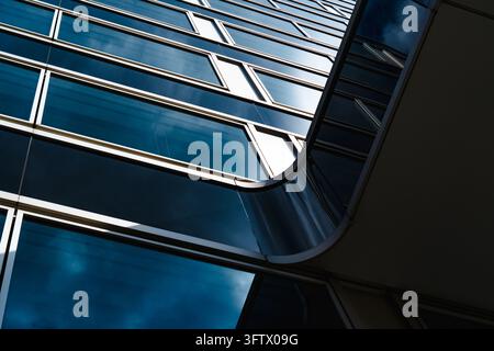 Eine Aufwärtsaufnahme zeigt blaue reflektierende Verglasung und silberne Fensterrahmen an einem modernen Bürogebäude. Eine weit geschwungene Kante unterbricht die Wiederholung Stockfoto
