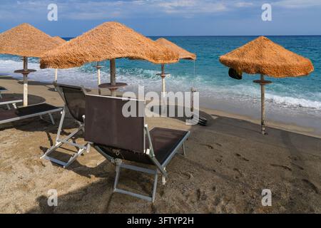 Sonnenliegen und Strohschirme auf sandigem Strand am türkisblauen Meer, Kalabrien, Italien Stockfoto