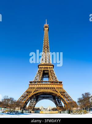 Eine niedrige Blickwinkelaufnahme des ikonischen Eiffelturms, umgeben von schneebedecktem Boden und Winterbäumen unter einem klaren blauen Himmel in Paris, Frankreich. Stockfoto