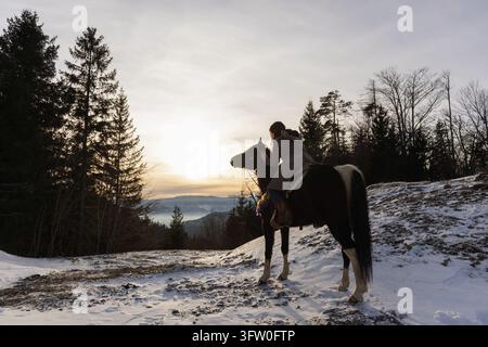 Beim Sonnenuntergang vom Pferderücken aus betrachtet, eine Frau, die einen friedlichen Moment umgeben von panormischen Bergaussichten und winterlicher Natur genießt. Stockfoto