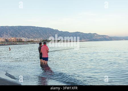 Albanien, Velipoja 12 09 2025: Zwei Männer angeln mit Ruten im Meerwasser am Strand von Velipoja mit Hotelgebäuden und Bergkulisse. Stockfoto