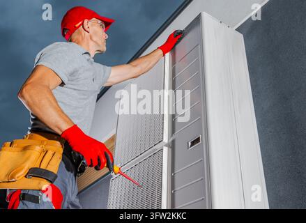 Niedriger Blick auf einen Techniker, der einen Werkzeuggürtel trägt und einen Schraubenzieher hält, während er das obere Gehäuse einer Außenwärmepumpe überprüft. Stockfoto