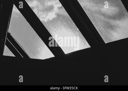 Eine schwarz-weiße Aufwärtsaufnahme einer Glasdachkonstruktion. Dunkle Metallträger bilden rauten- und trapezförmige Öffnungen. Das Glas ist nass und mit Regentropfen bedeckt Stockfoto