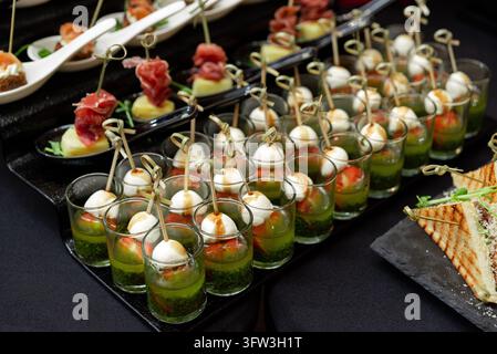 Caprese-Canapees mit Mozzarella-Kugeln und Kirschtomaten, die auf Spießchen in Schnapsgläsern serviert werden, die mit grüner Pesto-Sauce gefüllt sind, bei einem caterten Buffet. Stockfoto