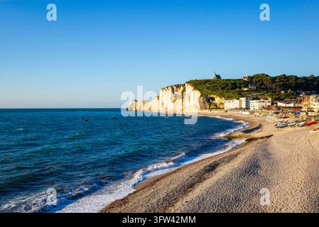 Eine weite Küstenansicht zeigt die Klippen, den Kiesstrand und die Küstenstadt Etretat im warmen Abendlicht, mit Booten, die am Ufer des Engländers angeschwemmt sind Stockfoto