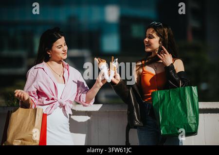 Zwei junge Frauen teilen sich Eisbecher, während sie im Freien mit Einkaufstüten stehen. Sie lächeln und plaudern zusammen in einer lockeren, sonnigen städtischen Umgebung. Stockfoto