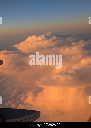 Aufragende Kumuluswolken, die von der untergehenden Sonne orange erleuchtet werden, gesehen von einem Flugzeugfenster mit der Flügelspitze am Rand des Bildes. Stockfoto