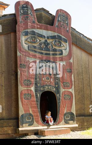 Ein Kind sitzt in der geschnitzten Tür des Stammeshauses von Chief Shakes auf Shakes Island in Wrangell, Alaska. Das Haus, eine Rekonstruktion des Civilian Conservation Corps aus dem Jahr 1940, wurde 2013 wiederaufgebaut und neu eingeweiht. Stockfoto
