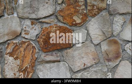 Mehrfarbige flache Fels- und Mörtelwand mit unterschiedlichen Steinformen und -größen, porösen rauen Oberflächen, natürlichen Kanten, rostiger und grauer Färbung Stockfoto