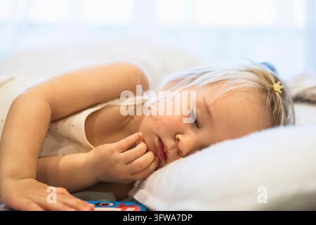 Ein blondes Kleinkind schläft, das Kind liegt friedlich auf einem weißen Kissen, die Augen geschlossen, in einem sanft beleuchteten Schlafzimmer Stockfoto