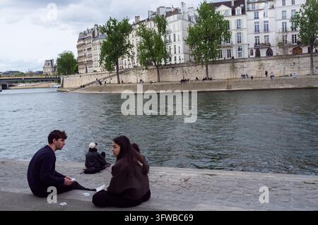 JUNGES PAAR IM GESPRÄCH AN DER SEINE, LINKE SEITE SONNTAG REPORTAGE, PARIS, FRANKREICH, 2026: Ein junger Mann und eine junge Frau unterhalten sich, während sie auf dem gepflasterten Ufer der Seine sitzen, mit anderen Menschen, die im Hintergrund entspannen, vor einer klassischen Pariser Stadtlandschaft. INFO: Die Linke Seite östlich von Notre-Dame vereint akademisches Erbe mit Flussromantik. Ein Spaziergang an historischen Universitäten, ruhigen botanischen Gärten und dem modernen Arabischen Weltinstitut vorbei zeigt eine lebendige, weniger touristische Ecke von Paris, die mit intellektueller Seele pulsiert. Stockfoto