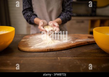 Hände formen frischen Teig auf bemehlter Holzplatte mit Rührschüssel in rustikaler Küche Stockfoto