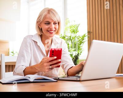 Selbstbewusste Frau, die mit einem Mobiltelefon und einem Laptop multitiert, während sie von zu Hause aus remote arbeitet. Stockfoto