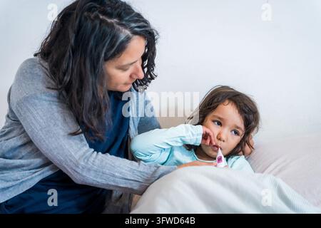 Kind mit Grippe im Bett mit Thermometer und Mutter zu Hause im Bett, Fürsorge und Liebe für die kleine Tochter. Stockfoto