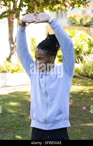 Afrikanischer Mann streckt sich mit erhobenen Armen auf dem Gartenrasen und zeigt ein weißes Armband und eine graue Kapuzenjacke Stockfoto