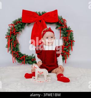 Nahaufnahme-Porträt eines süßen Mädchens in rotem Samtkleid mit rotem Kopfband, das auf dem Boden mit einem Holzschaukelpferd vor einem Weihnachtskranz sitzt Stockfoto