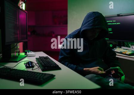 Geheimnisvolle Person in einem Hoodie, die an einem Schreibtisch mit mehreren Monitoren sitzt, die Code anzeigen. Dunkler Raum mit rosa und grünem Neonlicht erzeugt eine intensive Stimmung. Konzip Stockfoto