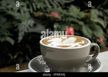 Eine frisch gebrühte Tasse heißen Cappuccino mit detaillierter geflügelter Pferde-Latte-Art, serviert auf einem Tisch mit üppigem grünen Blattgemüse-Hintergrund Stockfoto