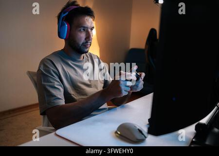 Junger männlicher Streamer mit bunten Kopfhörern, der den Controller festhält, konzentriert auf den Desktop-Bildschirm in einem dunklen Heim-Setup, während er live streamt Stockfoto