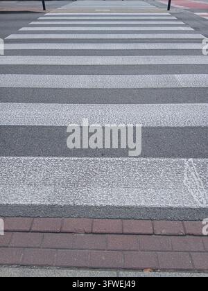 Eine detaillierte vertikale Ansicht einer weißen gestreiften Fußgängerüberquerung auf einem grauen Asphaltweg neben dem Gehweg. Stockfoto