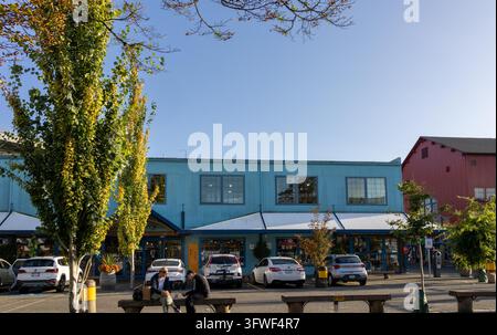 Vancouver, Kanada – 8. September 2026: Horizontale Straßenansicht von geparkten Autos, belaubten Bäumen und Fußgängern, die auf Bänken vor blauen Corrugas ruhen Stockfoto