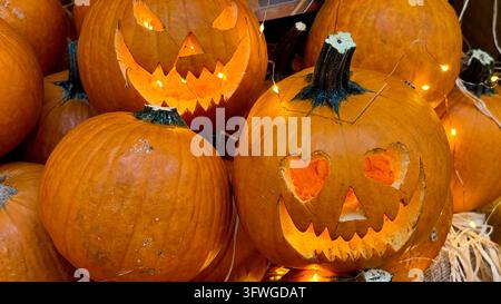 Nahaufnahme von geschnitzten Kürbislaternen, die mit warmem Licht leuchten, zwischen orangefarbenen Kürbissen und Stroh, Halloween-Dekoration. Stockfoto