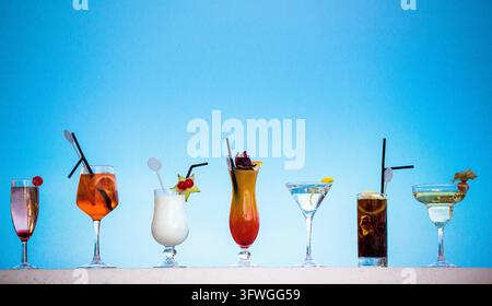 Eine Reihe von sieben verschiedenen Cocktails in verschiedenen Gläsern steht auf einer Kante vor einem klaren blauen Himmel. Stockfoto