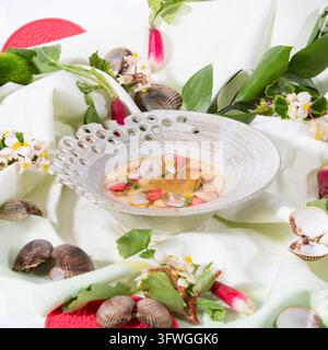 Eine rustikale Keramikschüssel gefüllt mit cremiger Suppe, die Muscheln, geschnittene Radieschen und essbare Blumen enthält, umgeben von frischen Zutaten auf einem weißen Tuch. Stockfoto
