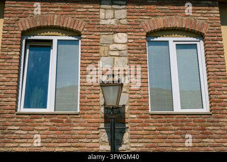 Strukturierte rote Backsteinfassade mit zwei weiß gerahmten Fenstern, horizontalen Jalousien, gewölbten Backsteinverzierungen, Steinakzenten und einer zentralen Eisenlaterne Stockfoto