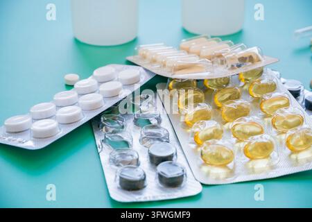 Weiße Tabletten, gelbe Weichgel-Kapseln und dunkle Pillen in sich überlappenden Blisterpackungen neben weißen Probenbehältern auf einer türkisfarbenen Oberfläche Stockfoto