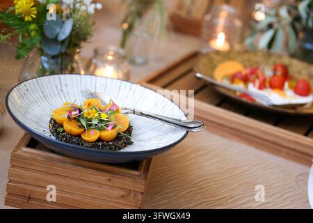 Eine Nahaufnahme eines angerichteten Gerichts mit Quinoa, gelben Tomaten und essbaren Blumen, serviert in einer gestreiften Keramikschale auf einem Holztablett. Stockfoto
