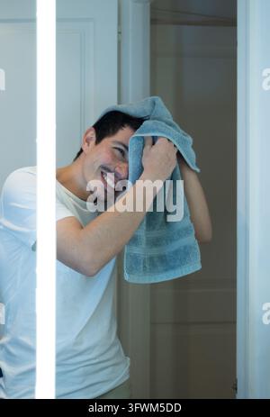 Vertikales Bild eines fröhlichen Mannes, der sich nach dem Duschen vor einem hellen Spiegel mit einem Handtuch die Haare trocknet Stockfoto