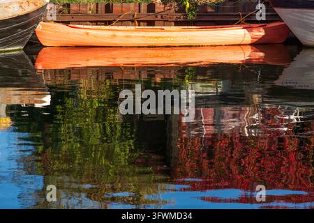 Ein lebhaftes rot-oranges Holz- oder Kompositkanu, das auf ruhigem Wasser festgemacht ist und eine scharfe, farbenfrohe Spiegelung im ruhigen Wasser erzeugt Stockfoto