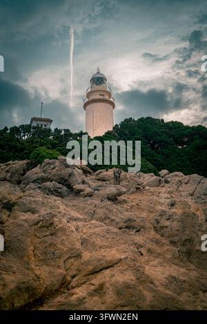 Der Leuchtturm von Cap de Formentor in Pollença, Spanien, erhebt sich markant vor einem Himmel voller dramatischer, dunkler Wolken. Im Vordergrund dominieren schroffe Stockfoto